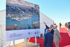 A group of white men in suits look at a billboard sign showing a port development