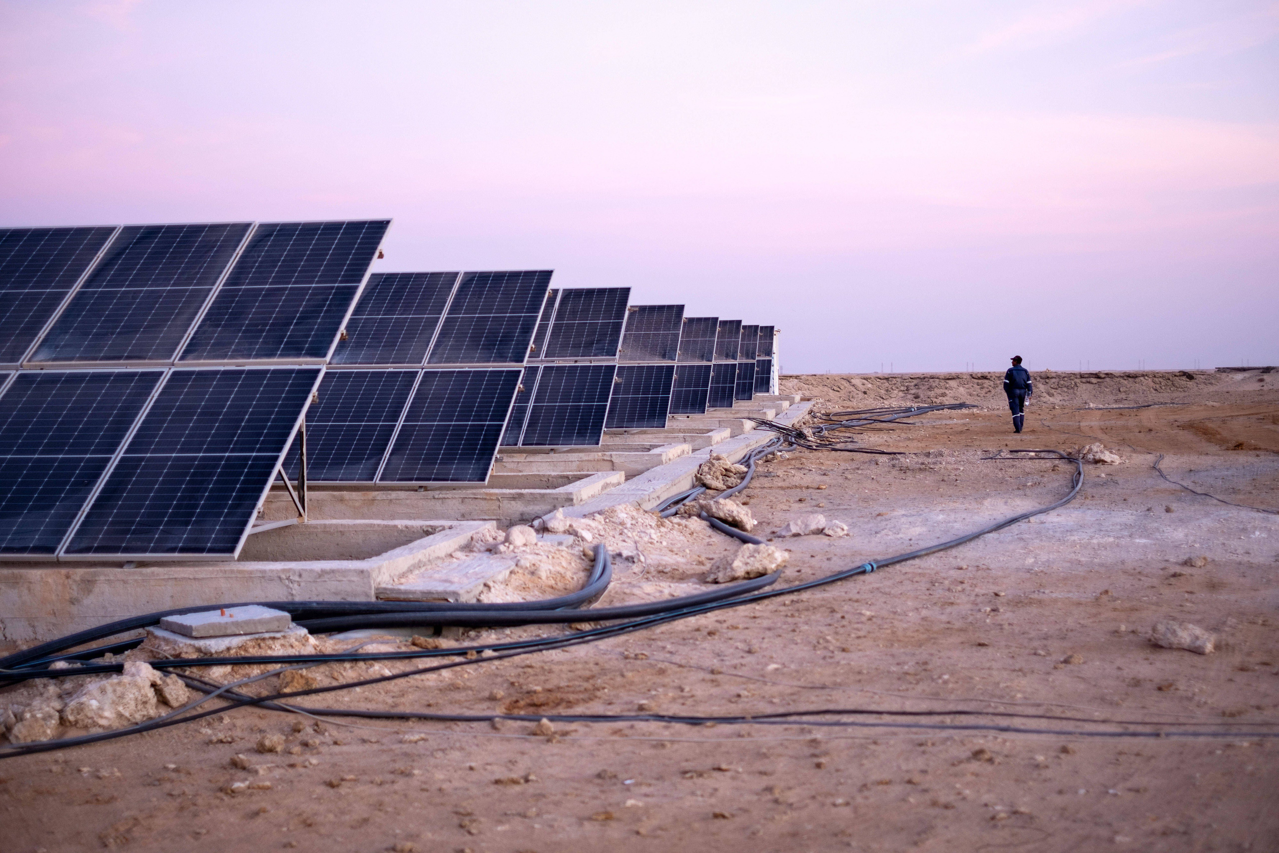 A man walks past solar panels and pipes in the desert